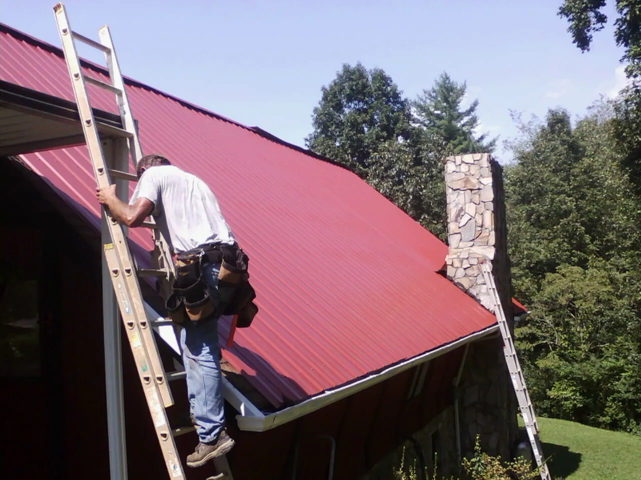 Roofer on a ladder beside a red metal roof and stone chimney — Soffit & Fascia Repair by Roofing Brothers in Coulee Dam